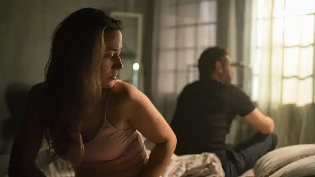 A woman and man sitting with their backs to each other and not talking while they sit on a bed.