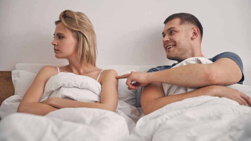 A man points at a woman’s shoulder while she sits in bed looking away.