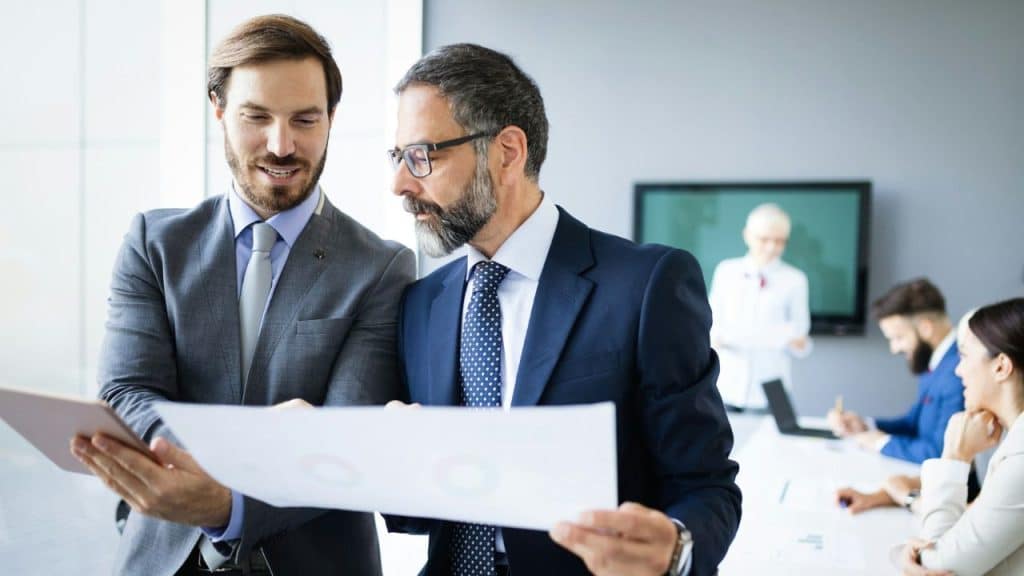 Two men in suits stand together looking at a document in a bright office.