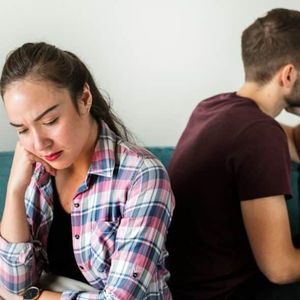A woman looking sad and sitting with her back to a man who also looks away while they sit on a couch.