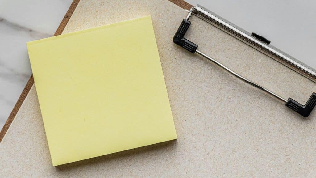 A blank yellow adhesive note is placed on top of a tan clipboard.