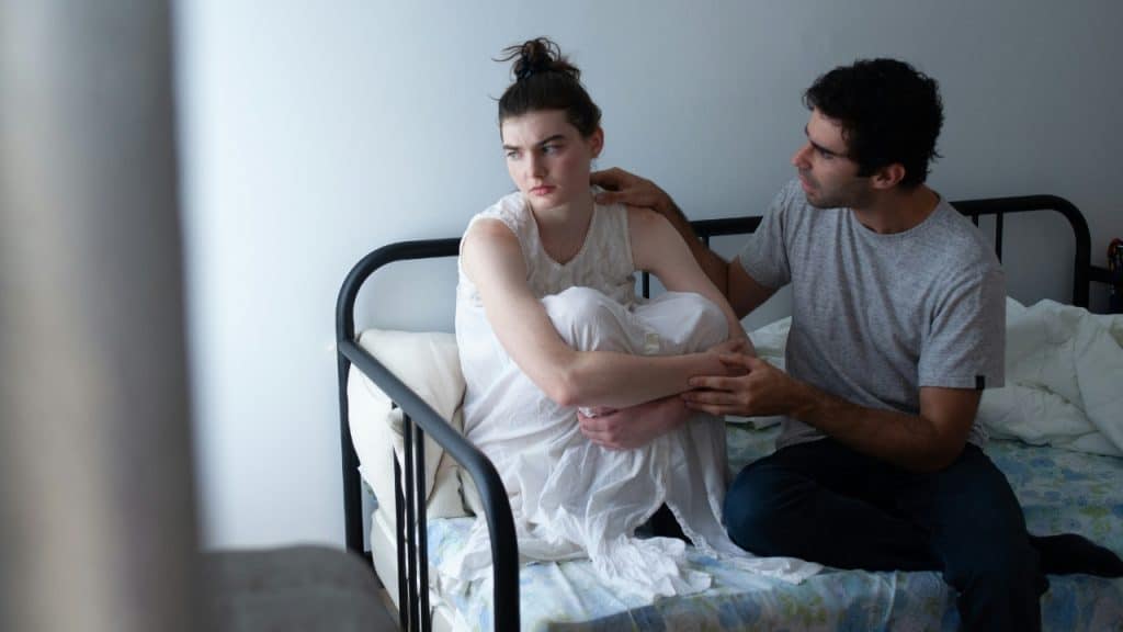 A man places a hand on the shoulder of a woman sitting curled up on a bed.