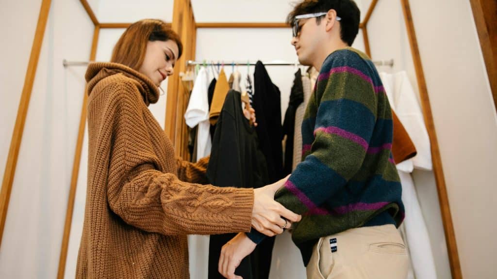 A woman in a brown sweater holds a man’s arm in a walk-in closet.