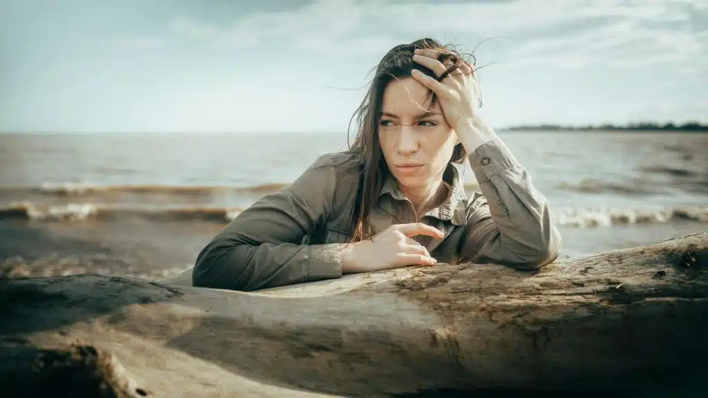 A woman resting on a driftwood log by the water, looking thoughtfully into the distance.