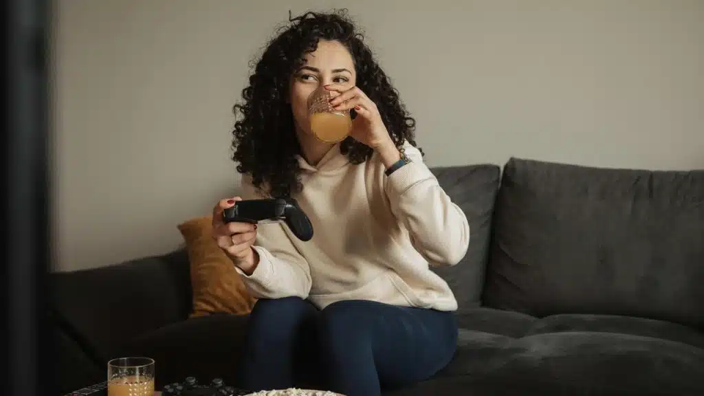 A woman drinks juice while holding a game controller on a couch.