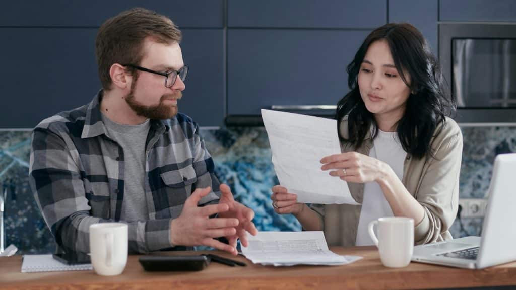 A man and woman sit at a table looking at papers with a laptop nearby.