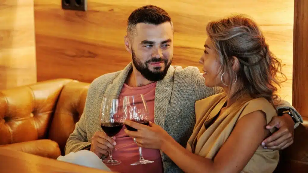 A man and woman sitting on a leather booth while holding glasses of red wine.