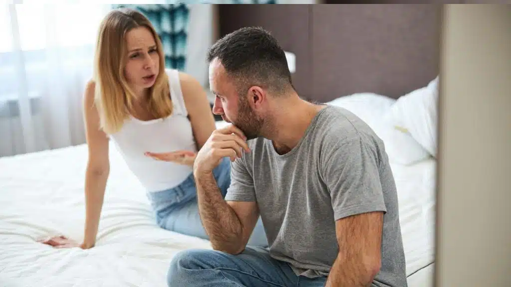 A displeased blonde lady is sitting on a bed and talking with her husband while the man is touching his chin.