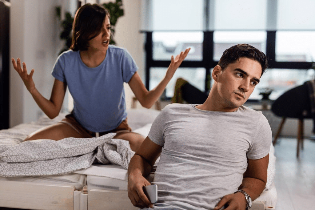 Young man sitting by the bed and ignoring his girlfriend