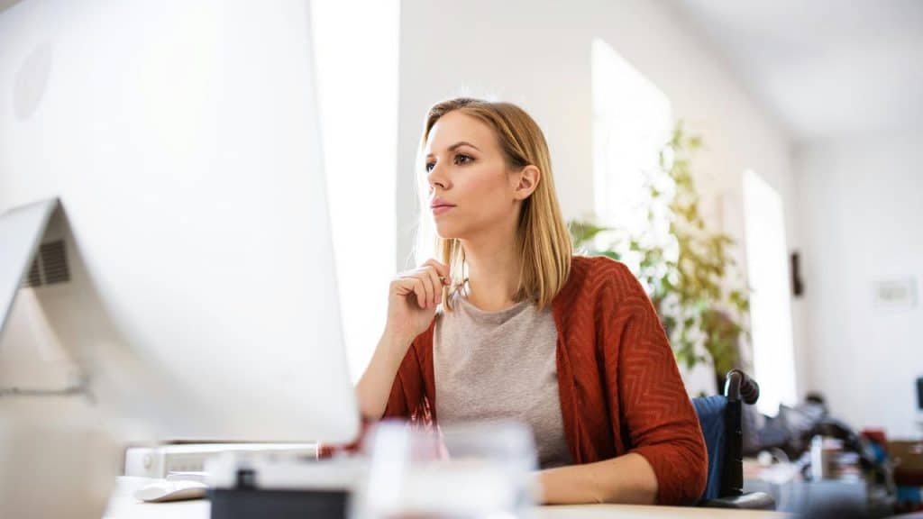 A woman sitting at a desk, thoughtfully looking at a computer screen.