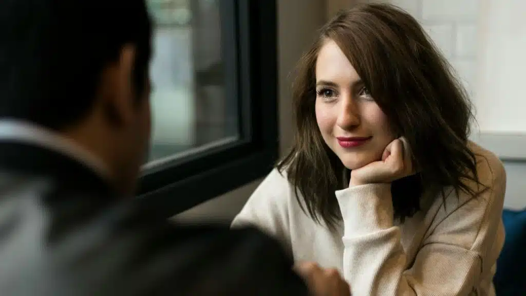 A woman with a slight smile sits across from a person, resting her chin on her hand.