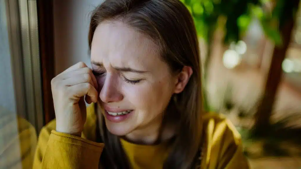 A distressed woman with light hair cries, wiping her eye with her hand.