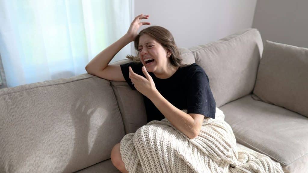 A young woman sits on a couch, wrapped in a blanket, with her hand on her head, crying and laughing.