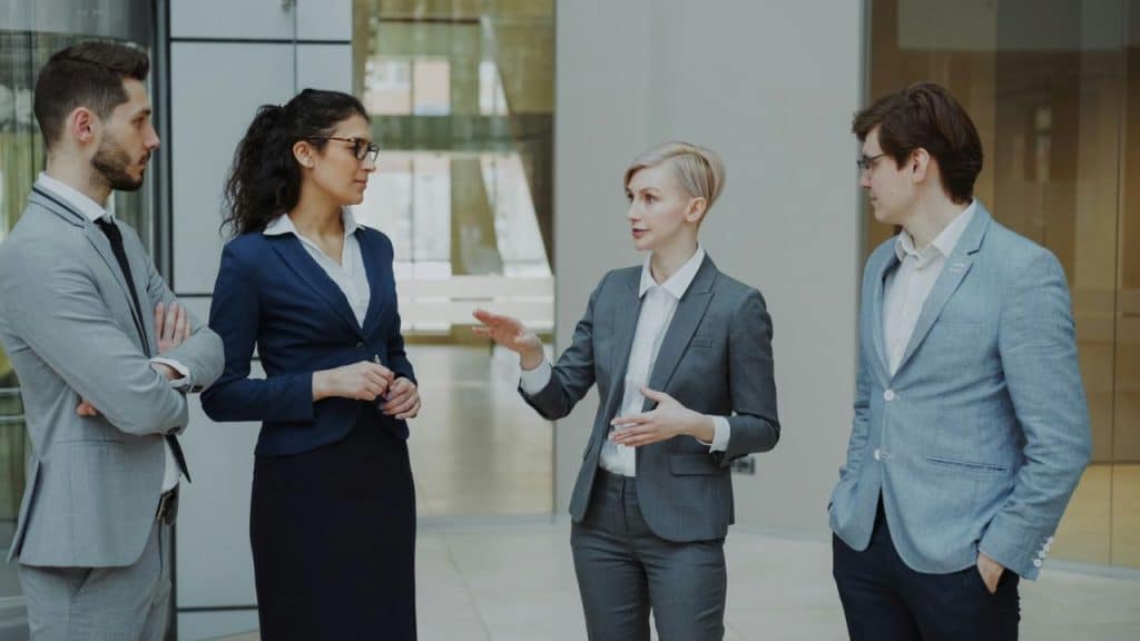 Four people in professional business attire standing and talking in a modern office space.
