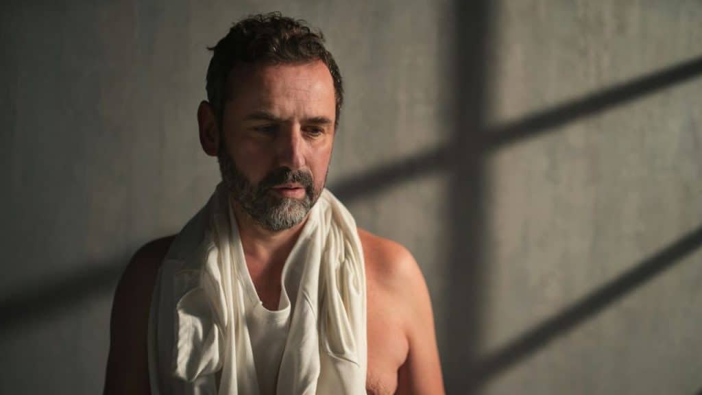 A man with a beard looks down, a white shirt draped over his shoulders.