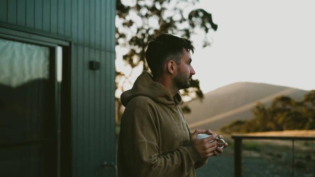 A man standing outdoors holding a cup and looking toward distant hills at sunset.