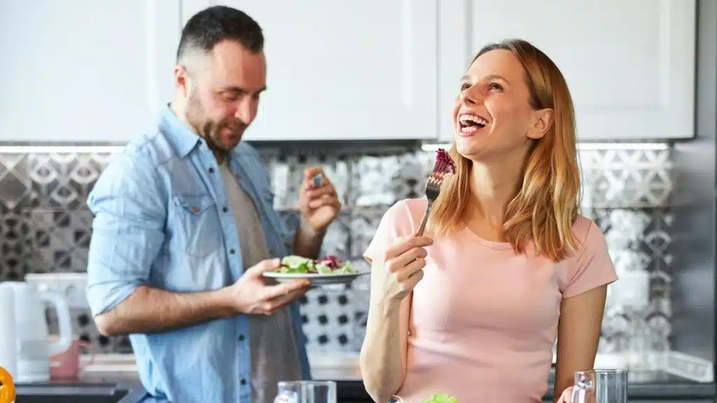 A woman laughs while holding a fork, while a man holds a plate behind her.