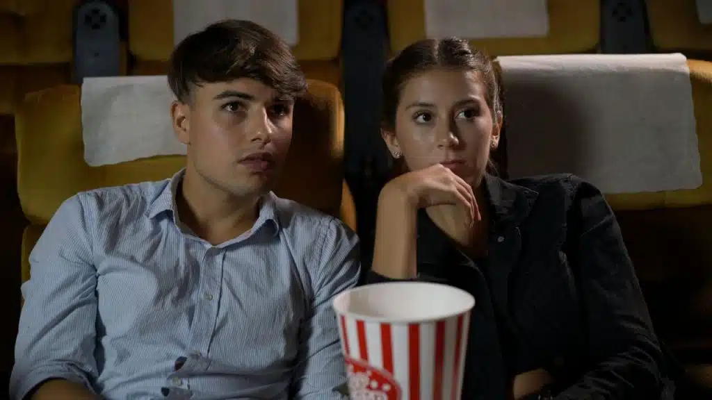 A man and woman sit in a dark theater with a popcorn bucket in front.