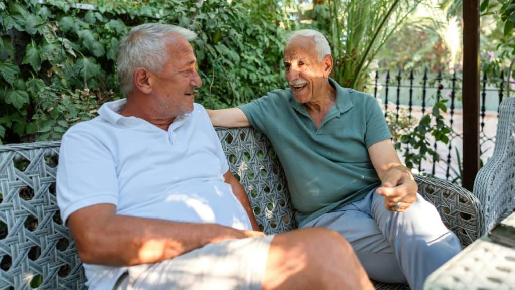 Two older men sit on an outdoor bench, smiling and talking in the sunlight.