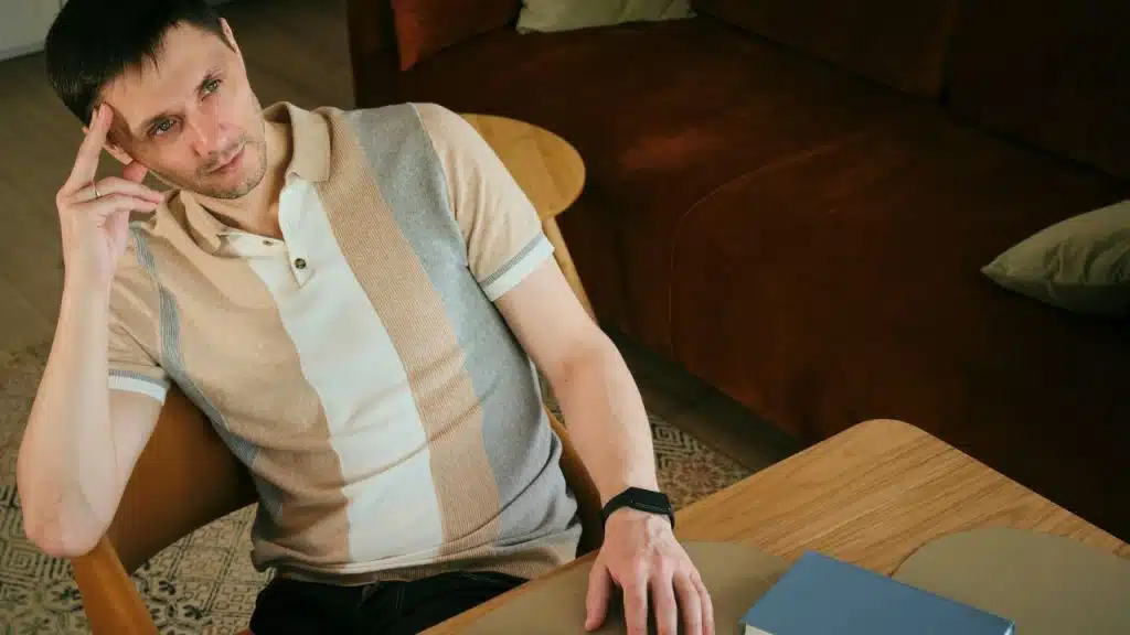 A man in a striped polo shirt sits at a table looking away thoughtfully.