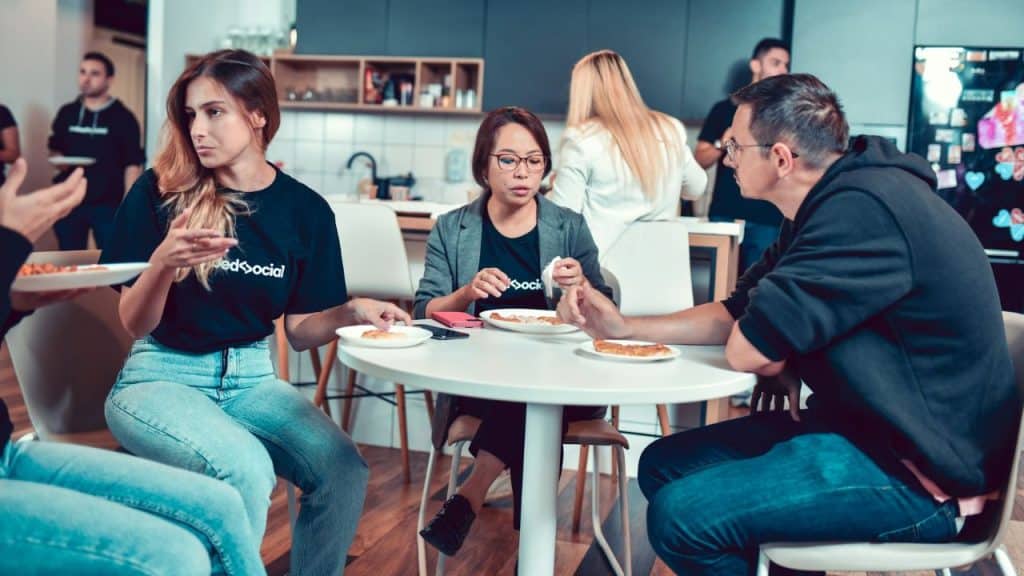 Three people sit at a round white table in a kitchen eating pizza and talking.