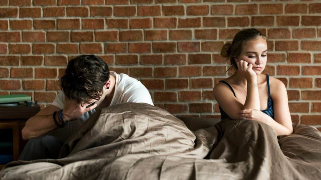 A man covers his face while a woman sits beside him looking away in bed.
