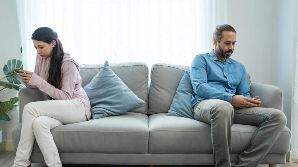 Two people sit at opposite ends of a grey couch, each looking down at a smartphone.