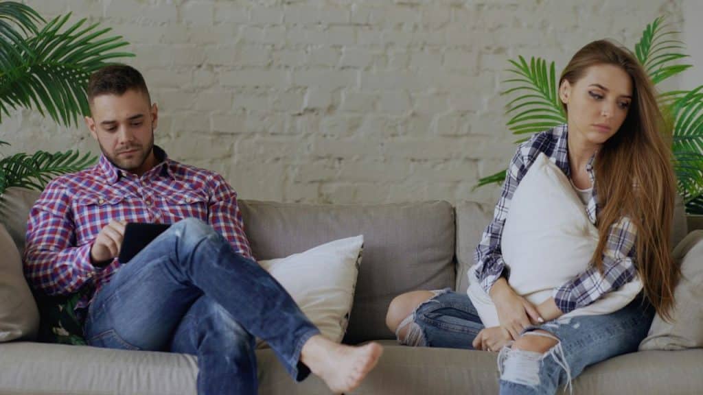 A man uses a tablet while a woman sits beside him holding a pillow looking away.