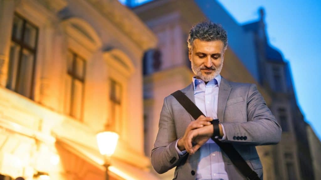 A man in a suit looks at his watch while standing outside at dusk.