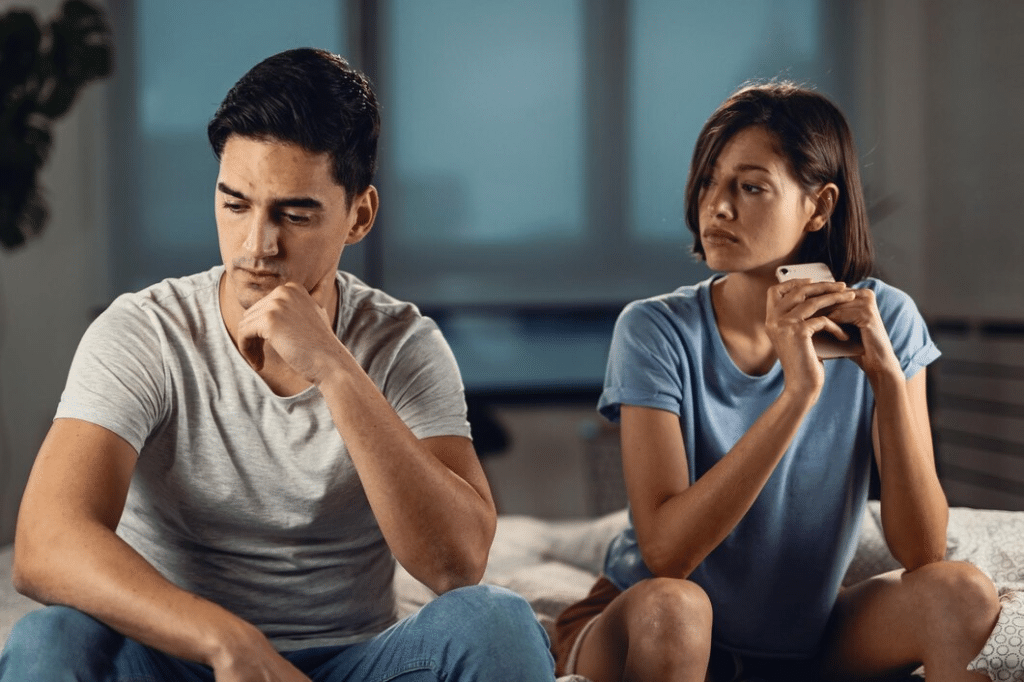 Man on bed ignoring pensive wife