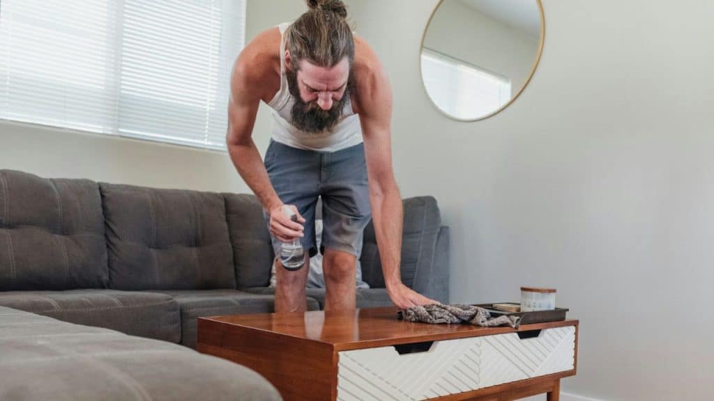 A bearded man in a white tank top uses a spray bottle to clean a table.