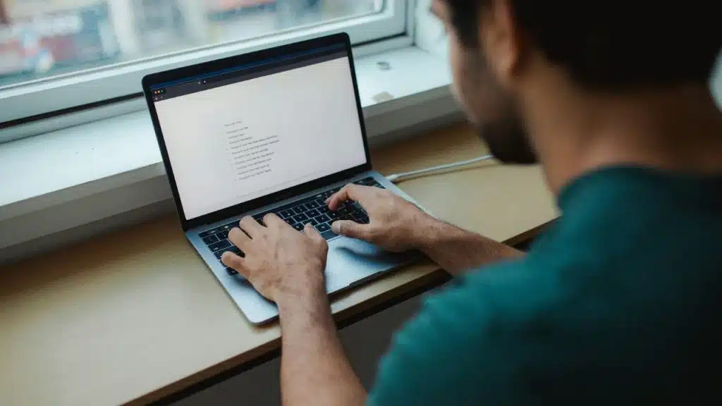 A man in a green shirt types on a laptop placed on a wooden desk.