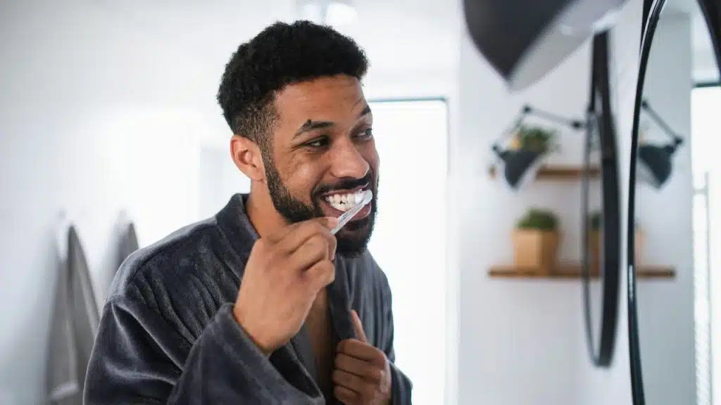 A smiling man in a grey robe brushes his teeth while looking into a bathroom mirror.