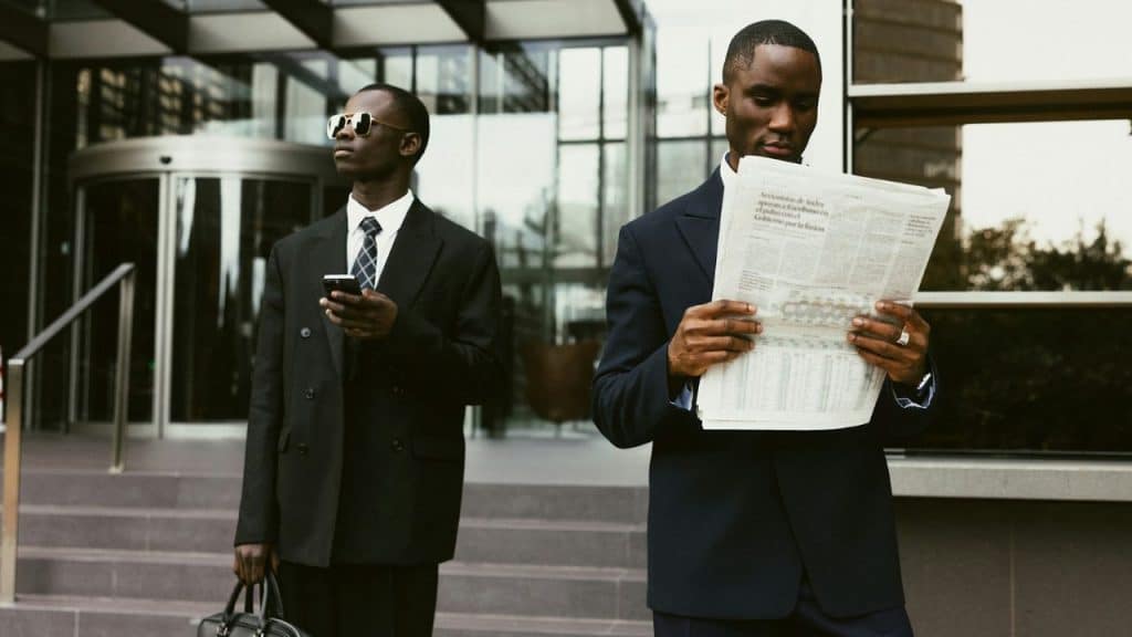 Two men in suits stand in an office hallway and hold some papers.