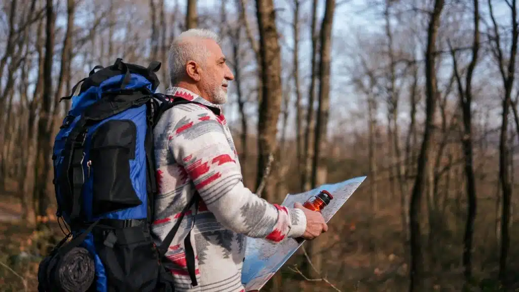 An older man with a backpack stands in a forest holding a map.