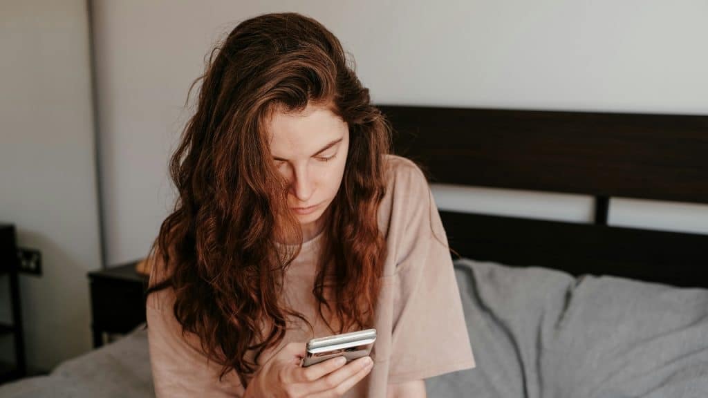 A woman with long wavy hair sits on a bed looking down at a smartphone.