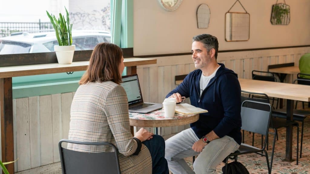 A man and woman sit at a small cafe table with a laptop.