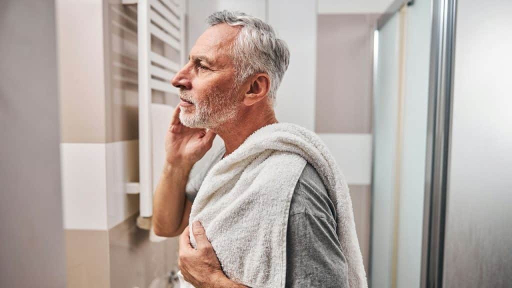 An older man with a towel over his shoulder looks at himself in a mirror.