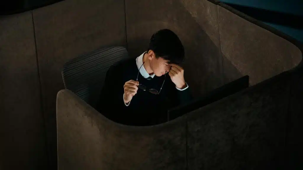 A man in a study booth rubbing his eyes while looking at a laptop.