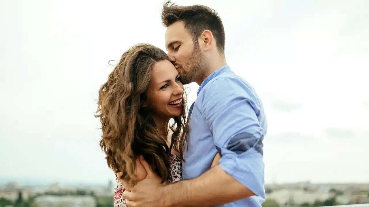 A couple embracing and smiling outdoors under a bright sky.