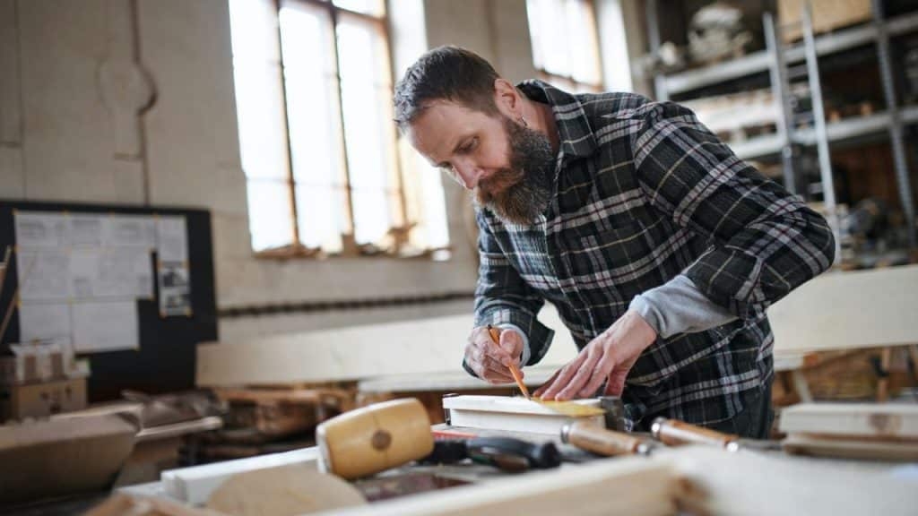 A bearded man in a plaid shirt uses a pencil and square to mark a piece of wood.