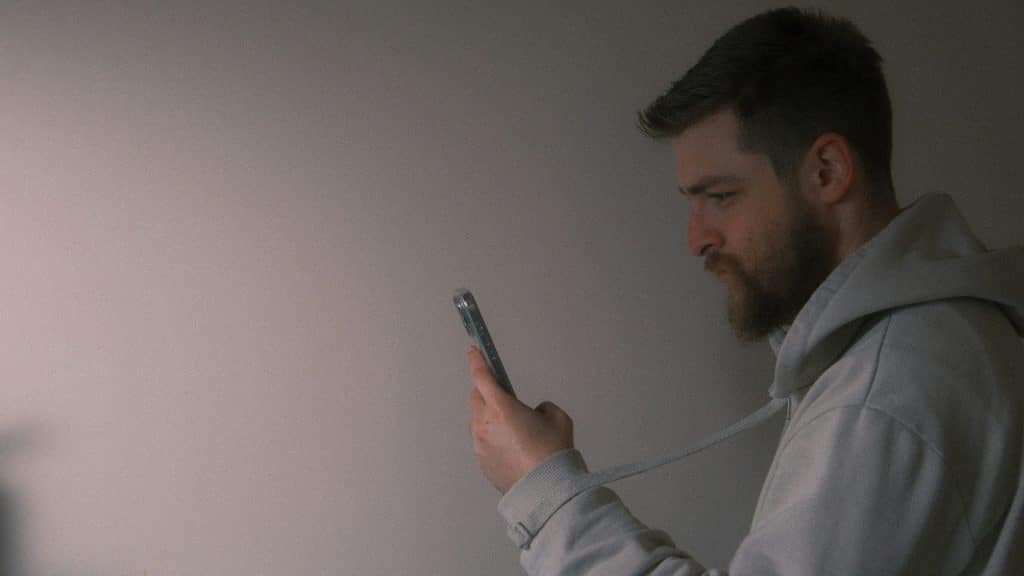 A man with a beard looks down at his smartphone against a plain background.
