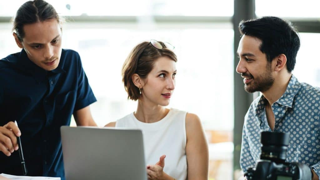 Three people sit around a laptop, with one woman looking toward a smiling man on her right.