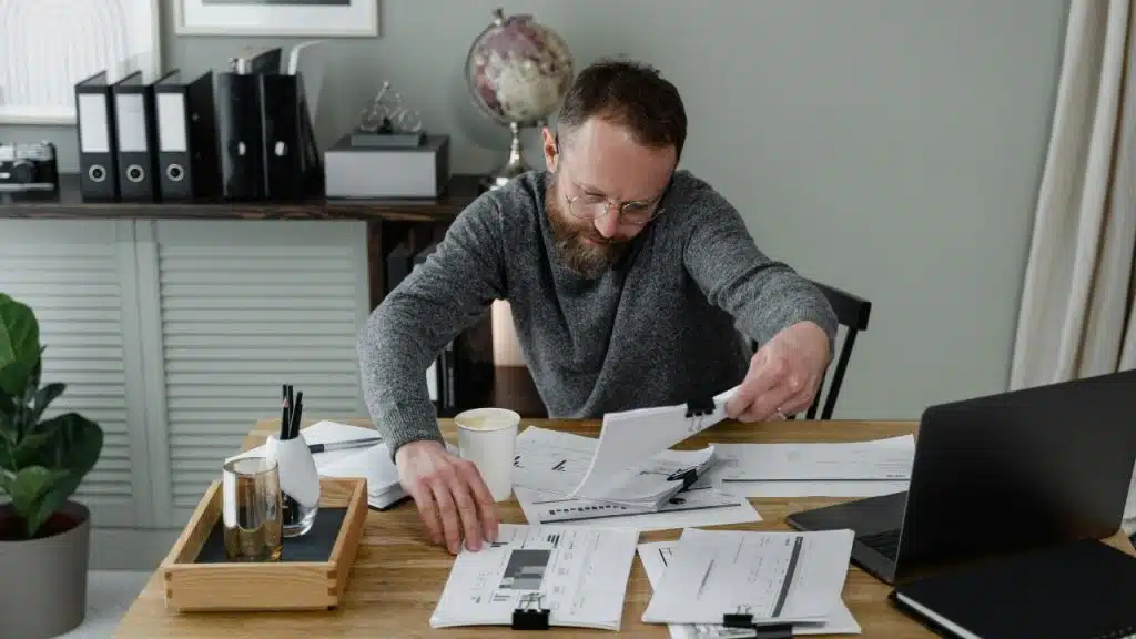 A man with a beard reviews documents and uses a phone at a desk.