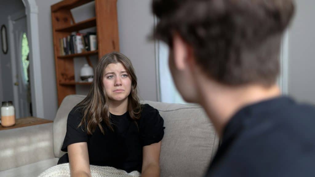 A woman with a sad expression sits on a sofa, looking at a blurred person.