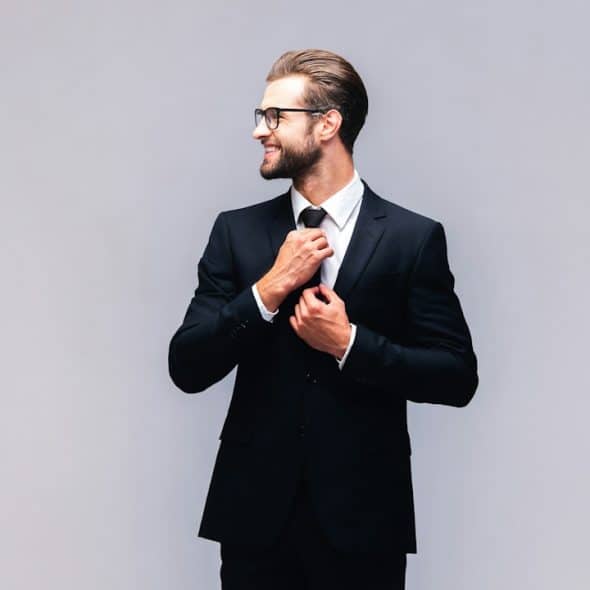 A lightly bearded man wearing glasses and a suit smiles while adjusting his tie.