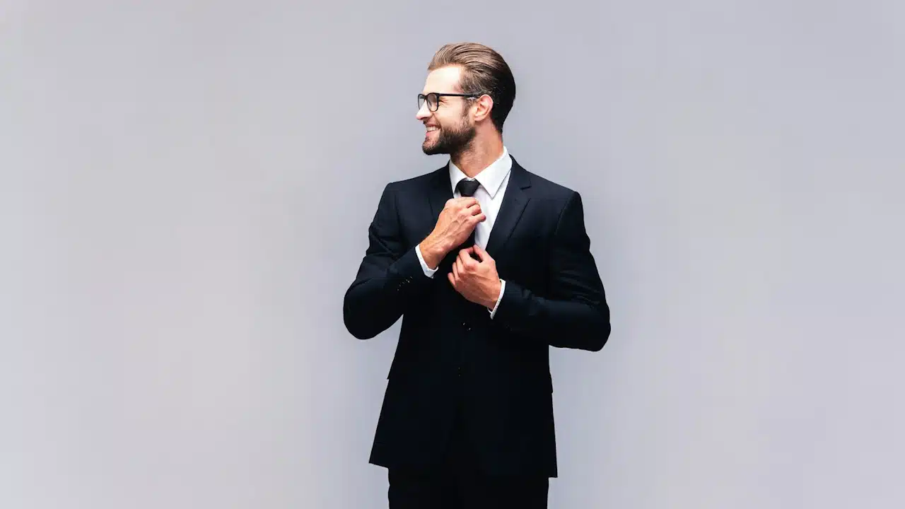 A lightly bearded man wearing glasses and a suit smiles while adjusting his tie.