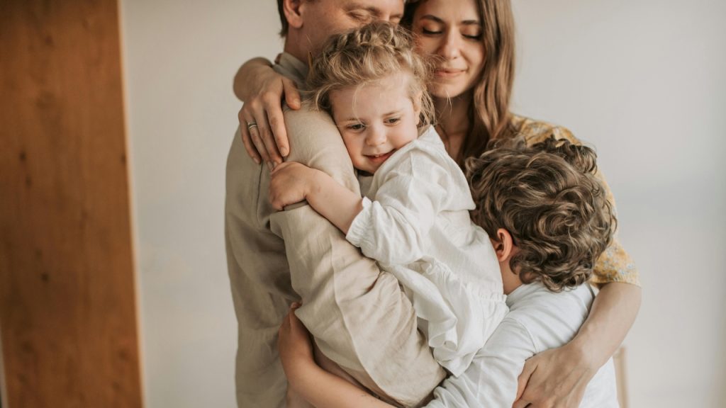 Two adults stand together while holding and hugging two young children in a close group.