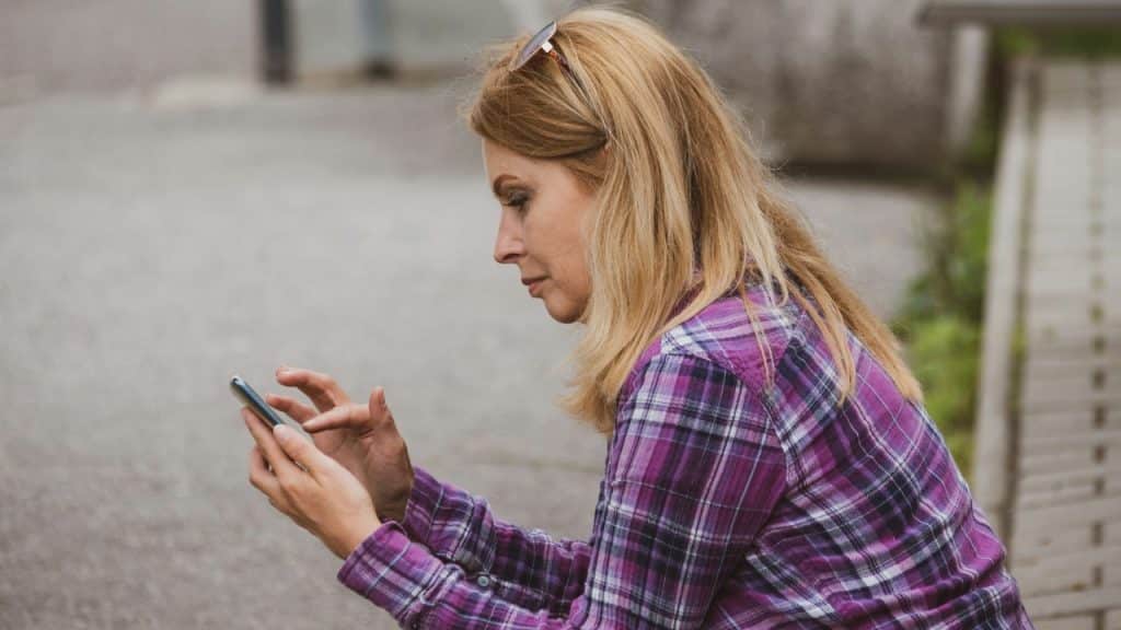 A woman with blonde hair in a plaid shirt looks down at her phone.