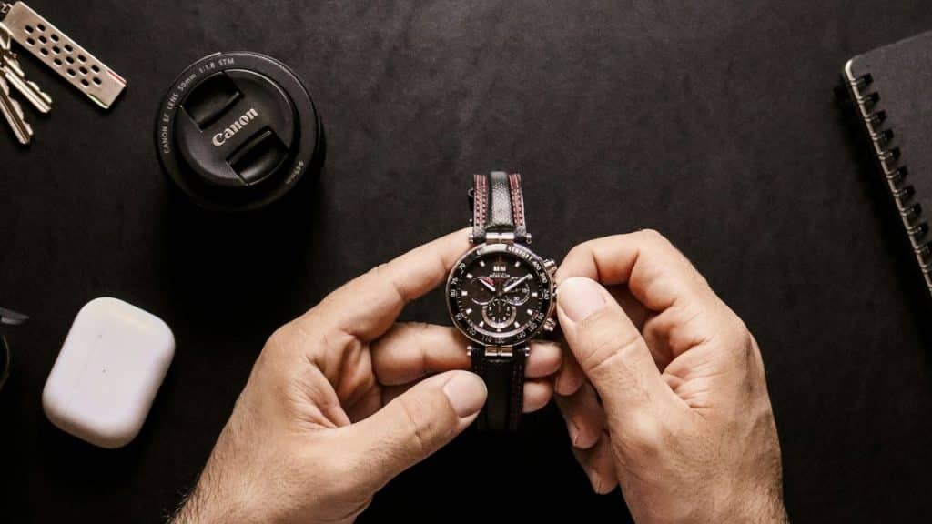 Hands adjust a wristwatch surrounded by a lens cap, keys, and a notebook.